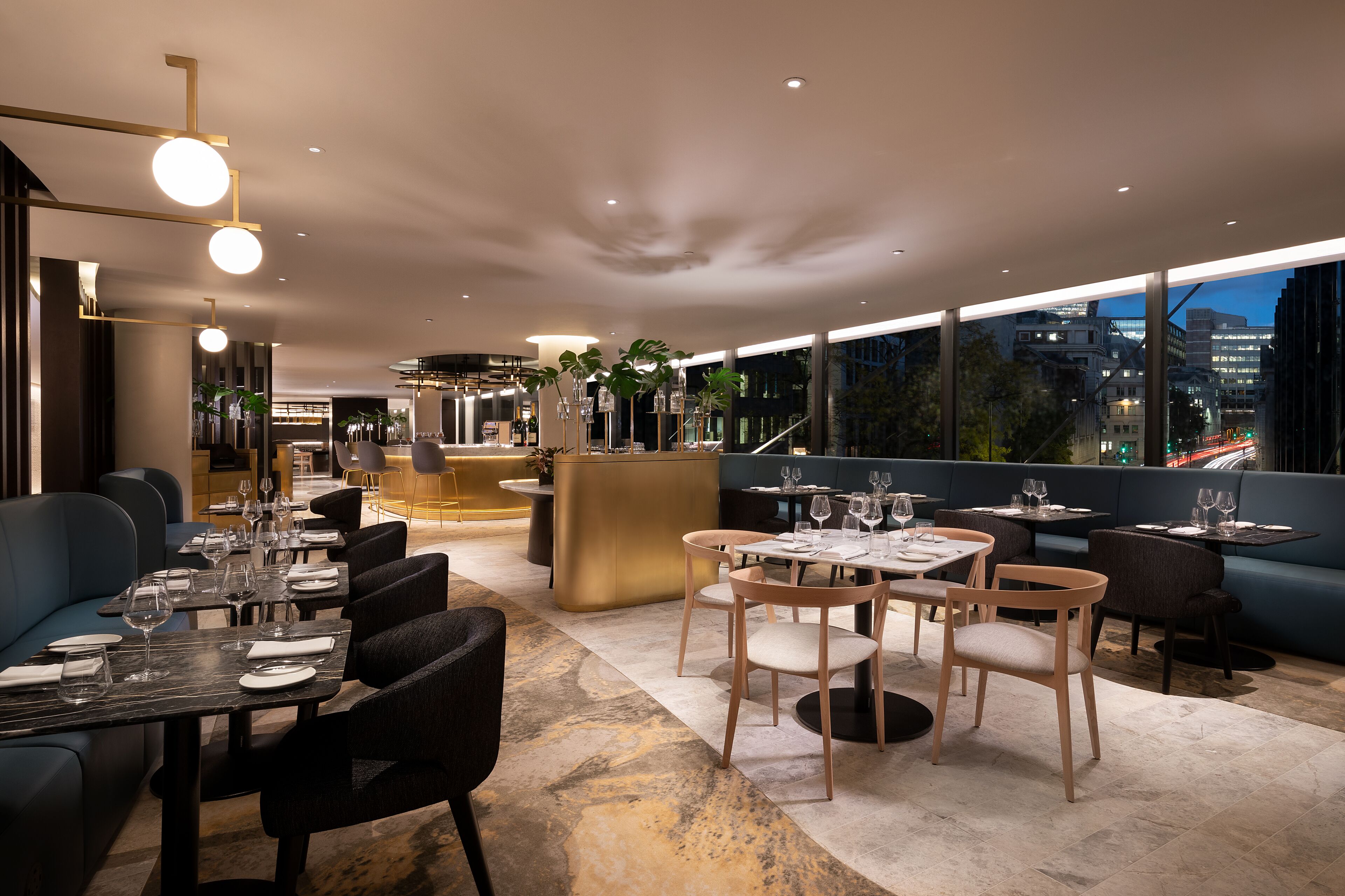 Un restaurant moderne avec des tables dressées pour le service, un bar et de grandes fenêtres offrant une vue sur la ville.