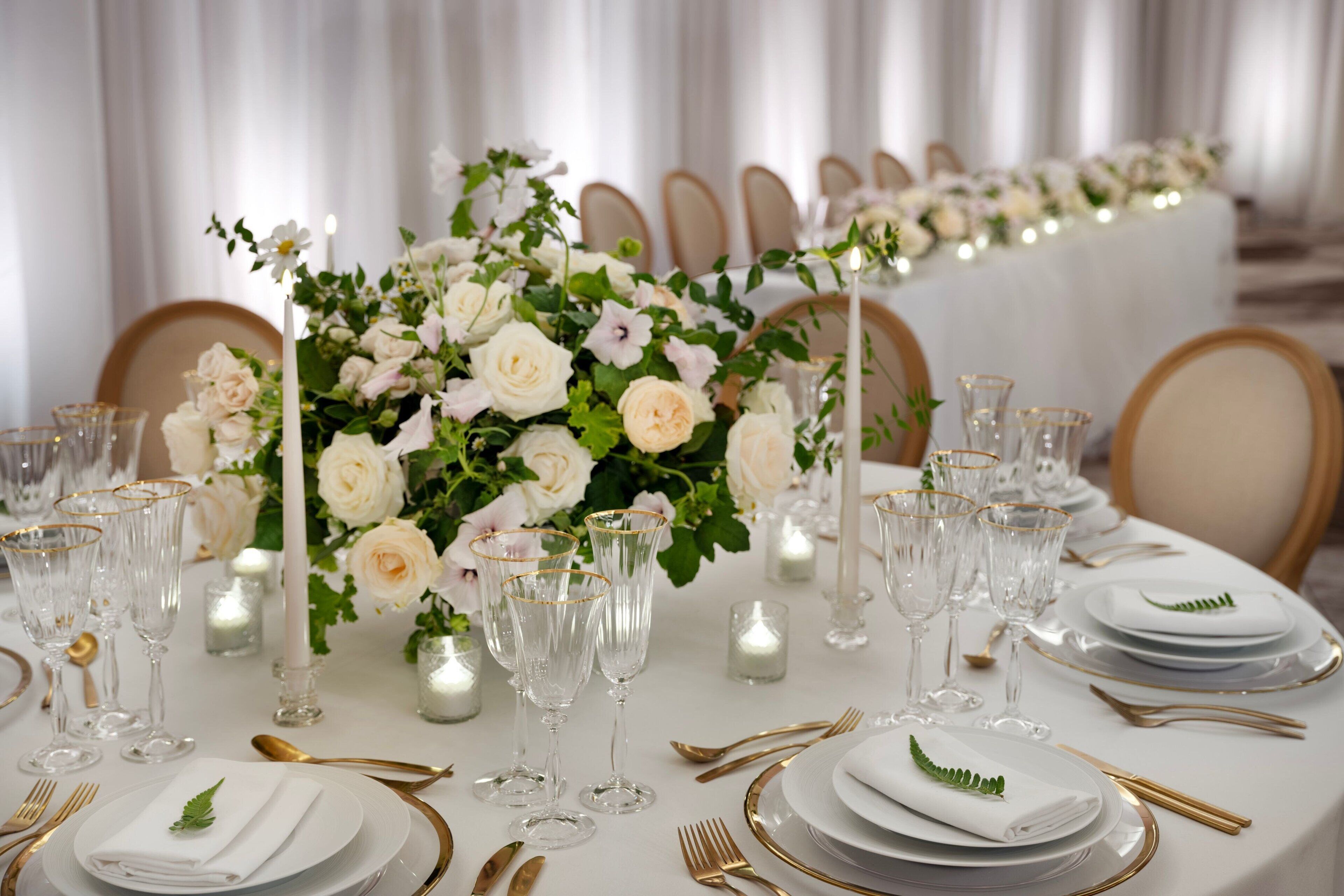 A formal dining table set with white plates, gold cutlery, and clear glassware, surrounded by white roses and greenery.