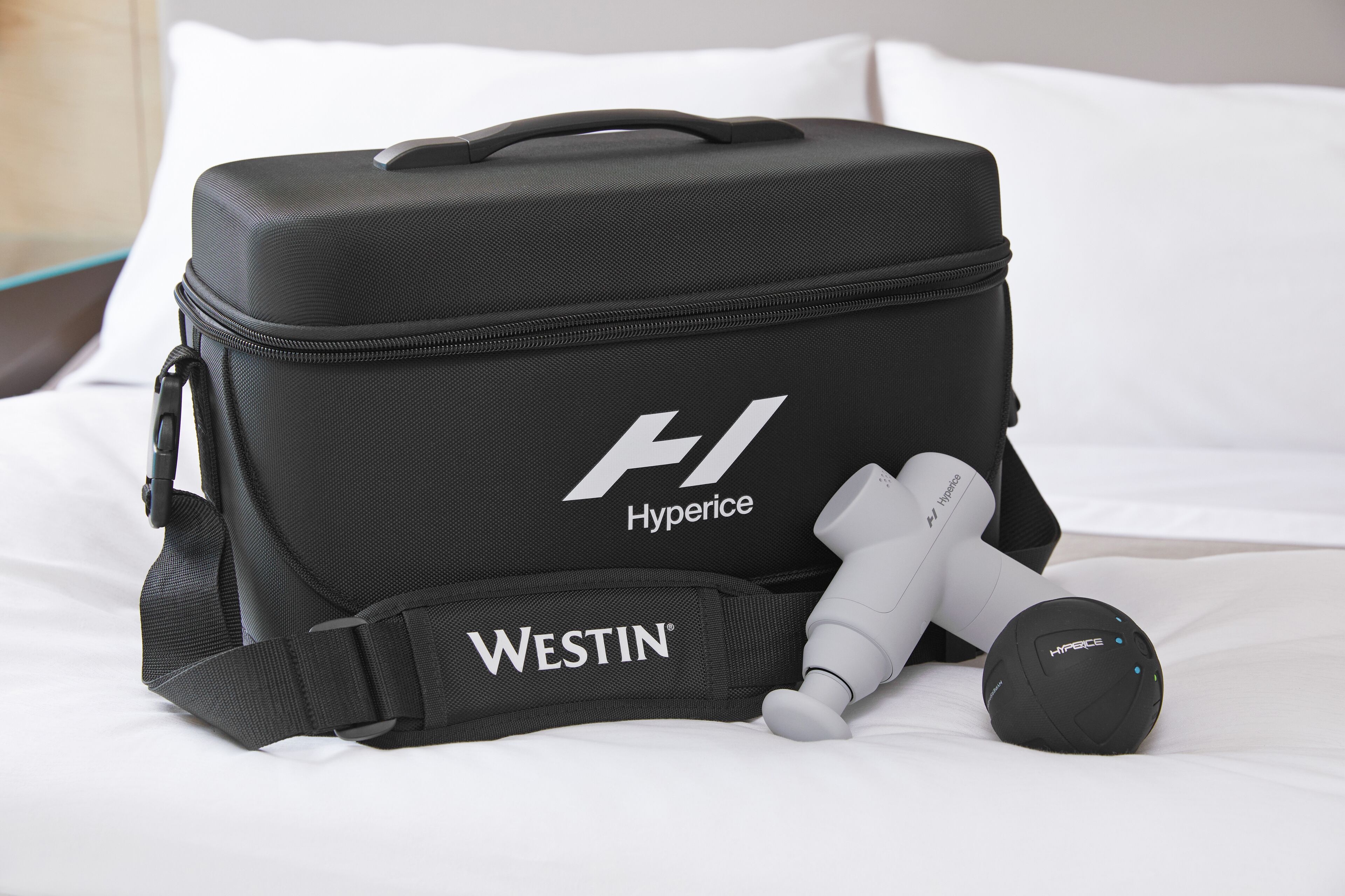 A black suitcase with "Hyperice" and "Westin" branding, a white pillow, and a white bedside table.