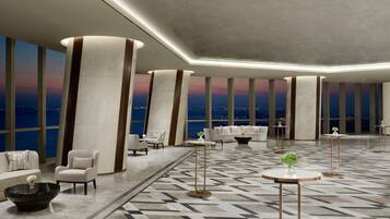 A spacious lobby with high ceilings, large columns, and a patterned floor. There are several seating areas with sofas and armchairs, and small tables with plants.