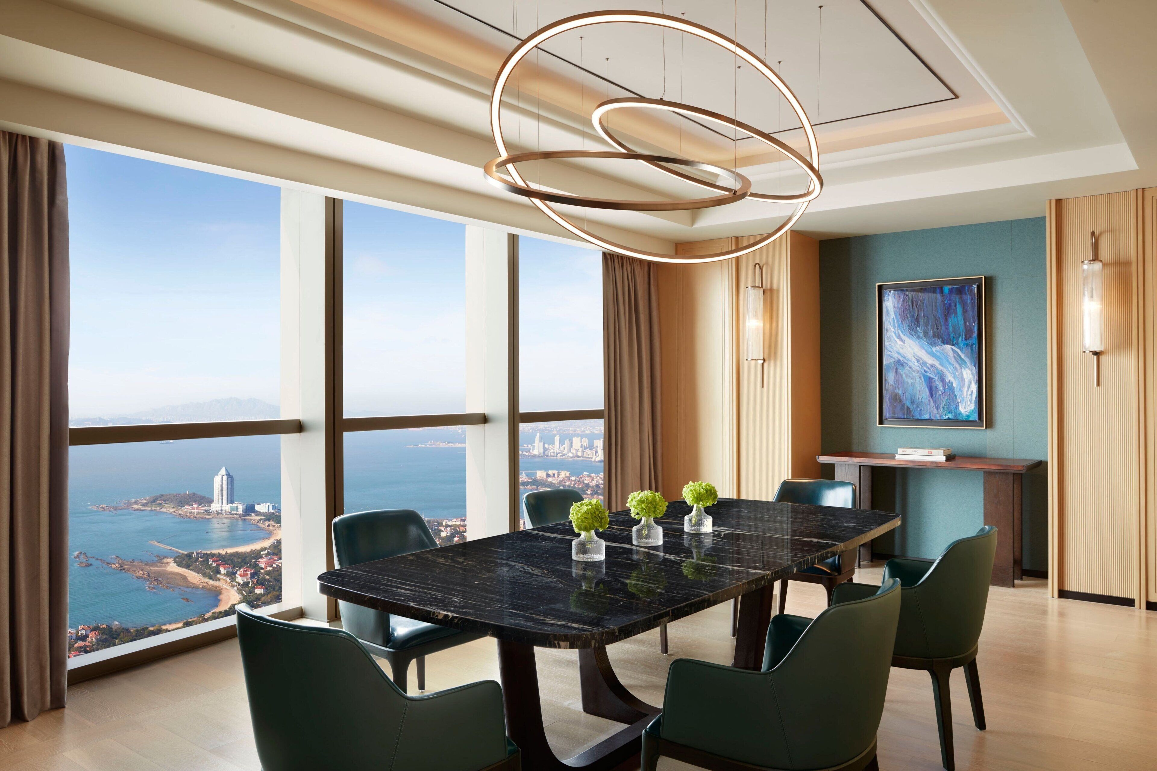 A modern dining room with a large table, green chairs, and a view of the sea and cityscape.