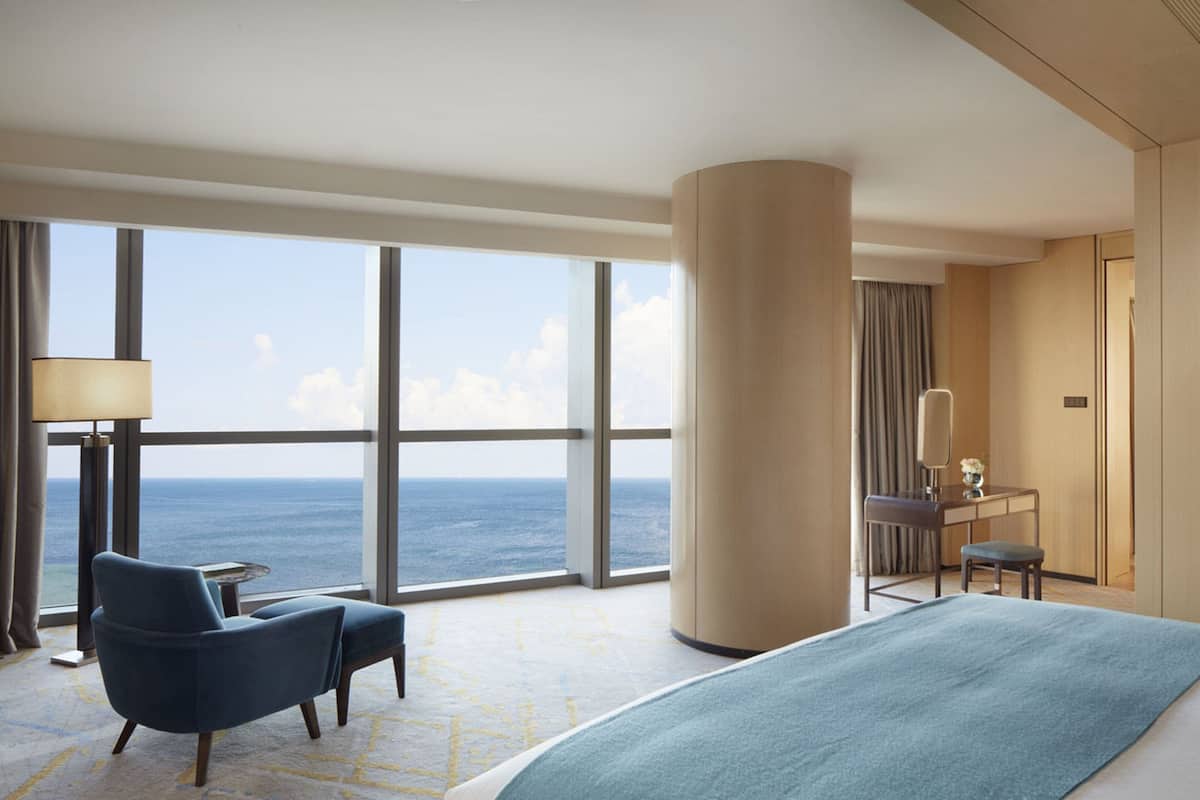 A hotel room with a large bed, a desk with a chair, a lamp, and a view of the ocean.