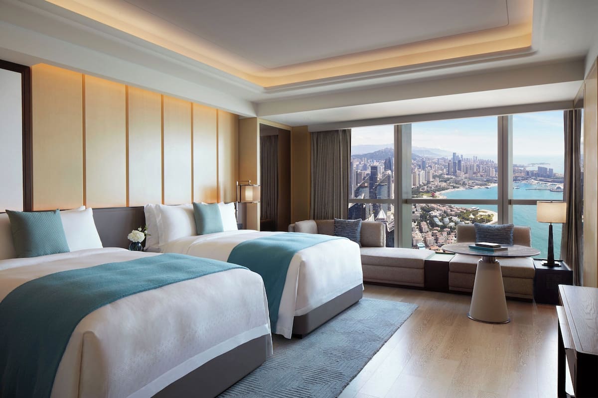 A hotel room with two beds, a sofa, a small table, and a large window offering a city view.