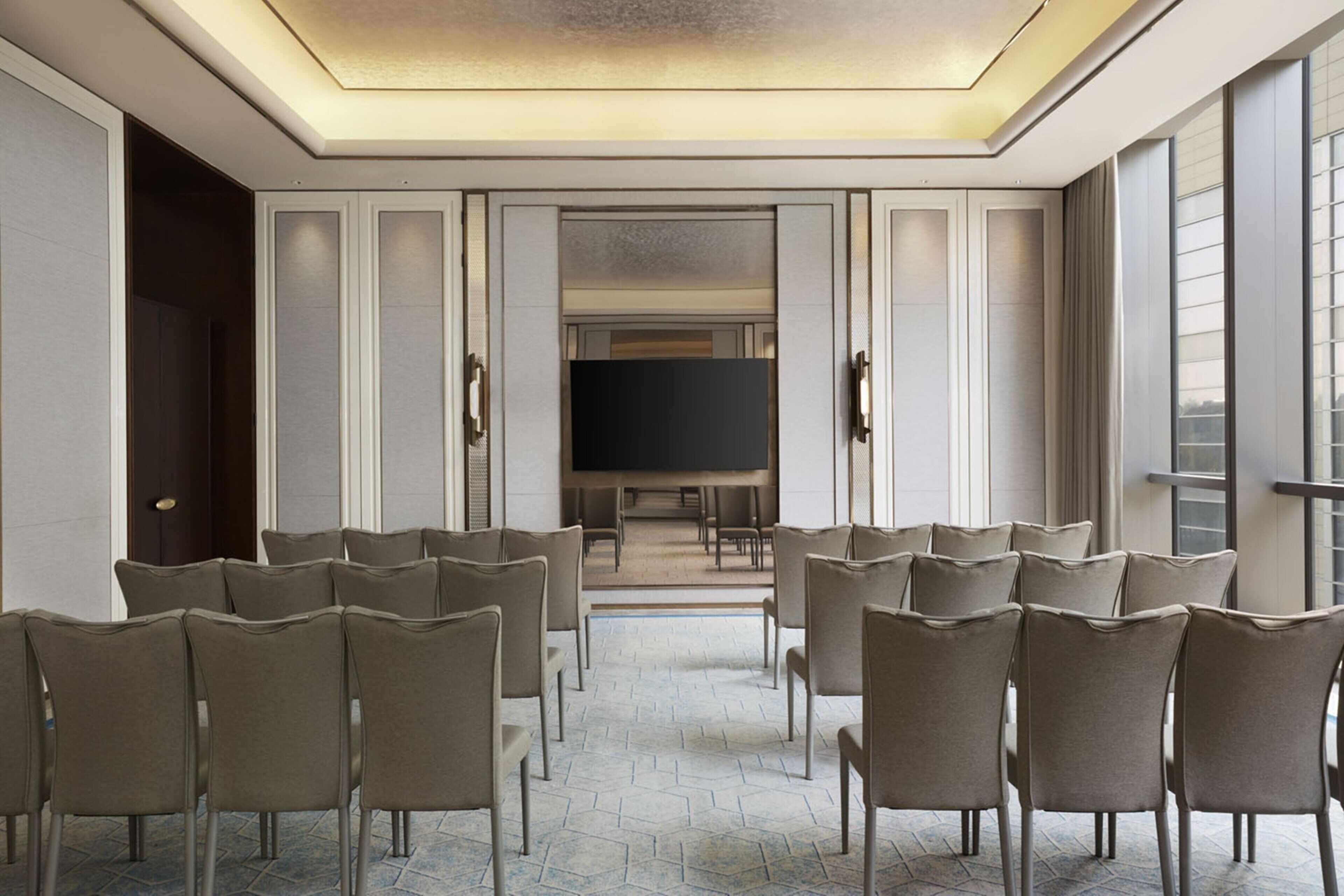 Un salón de conferencias con una pantalla grande, filas de sillas y vistas a través de una puerta abierta.