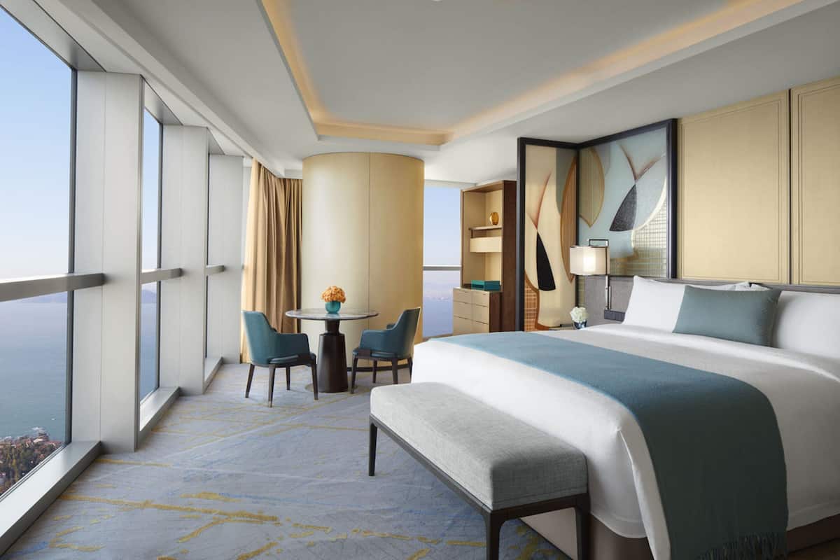 A hotel room with a large bed, a desk with chairs, a small table with a vase, and a view of the city through large windows.