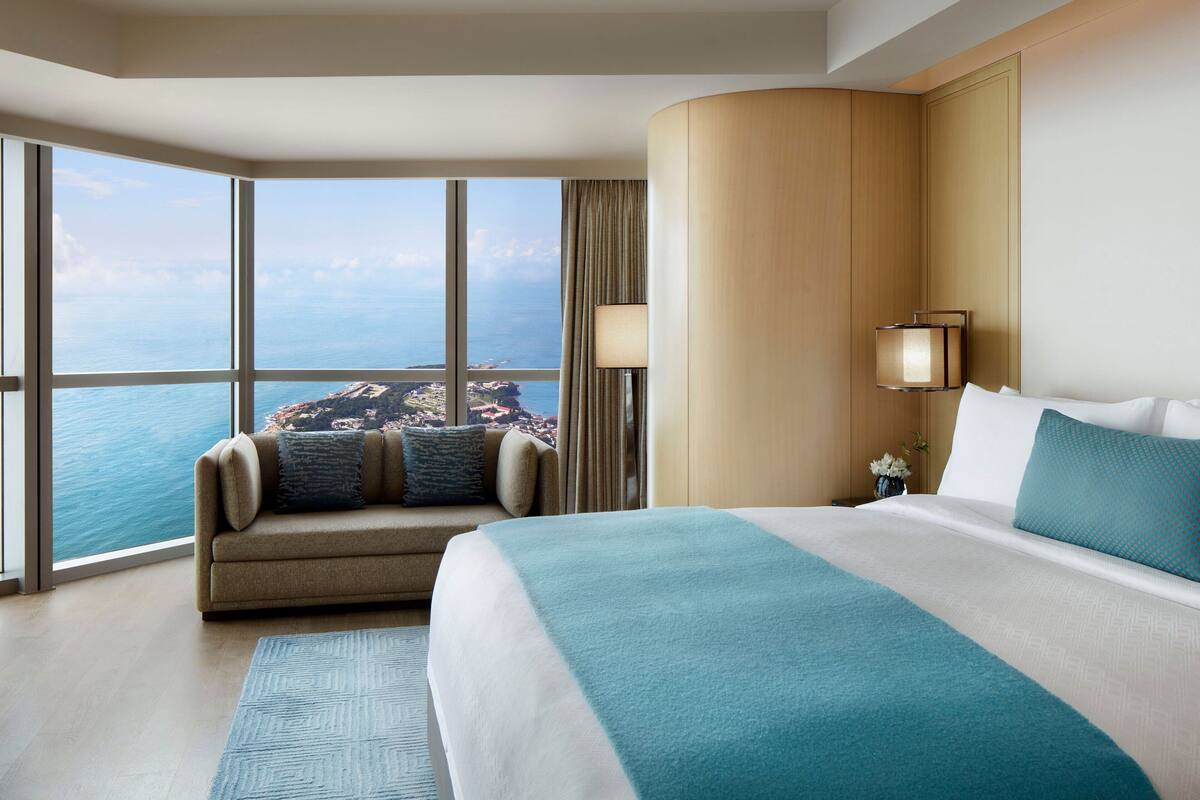 A hotel room with a large bed, a sofa, a view of the ocean, and a cityscape.