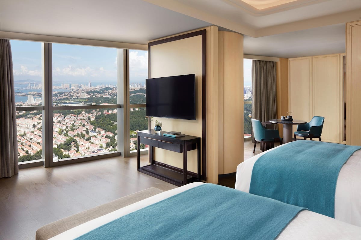 A hotel room with a large bed, a TV on a stand, a desk with a chair, and a view of a cityscape through large windows.