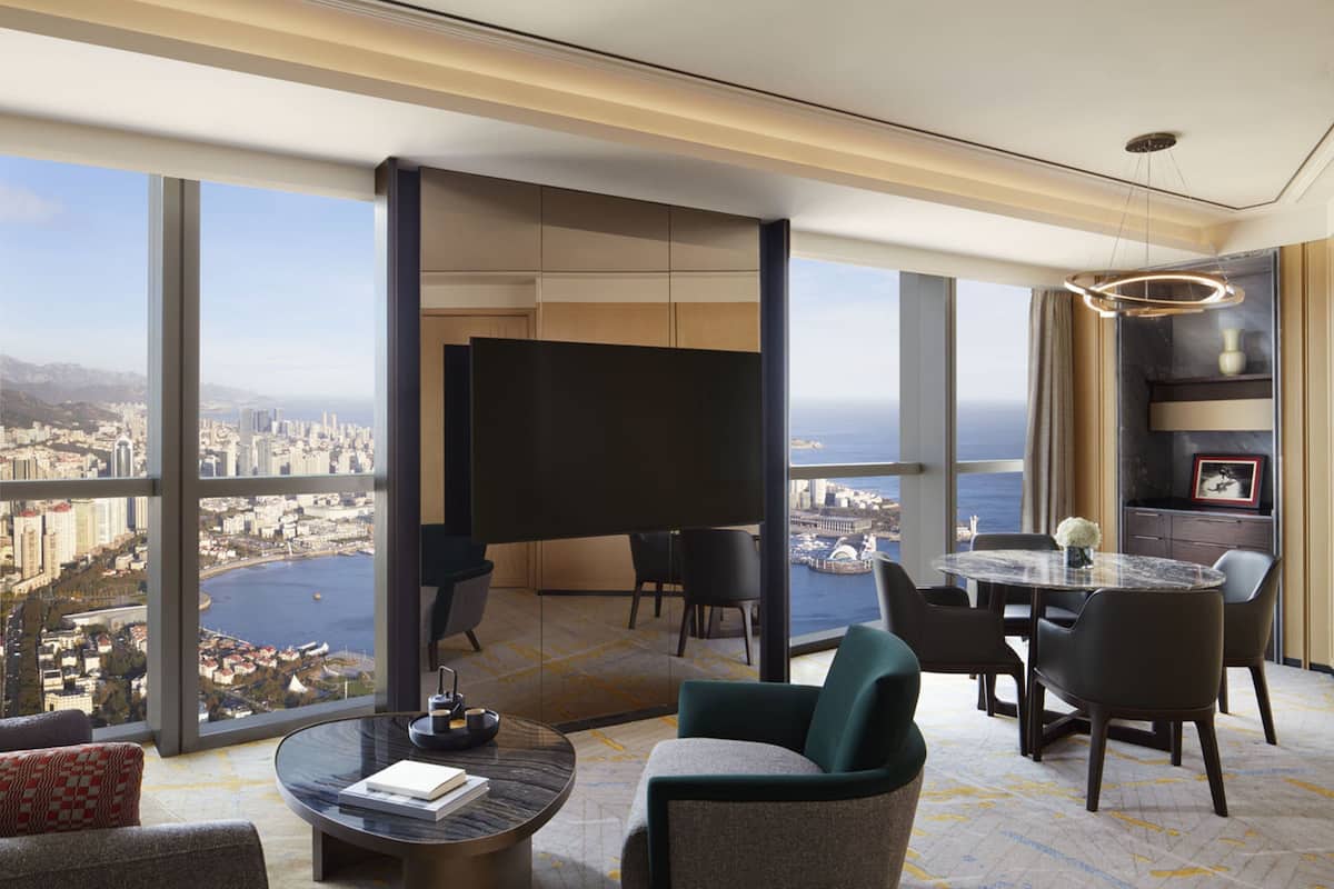 A modern hotel room with a panoramic city view, a dining area with a round table, and a seating arrangement with chairs.