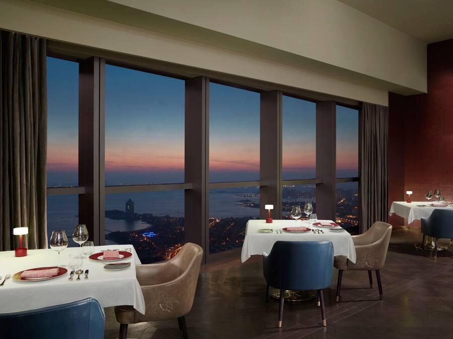 A dining area with tables set for two, large windows offering a city view at dusk, and a modern interior design.