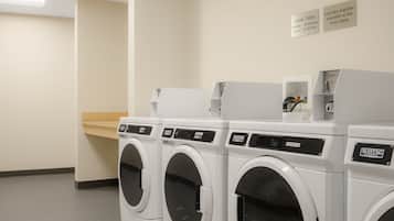 A laundry area with three front-loading washing machines, a dryer, and a wall-mounted ironing station. Signs indicate cycle times for the washer and dryer.