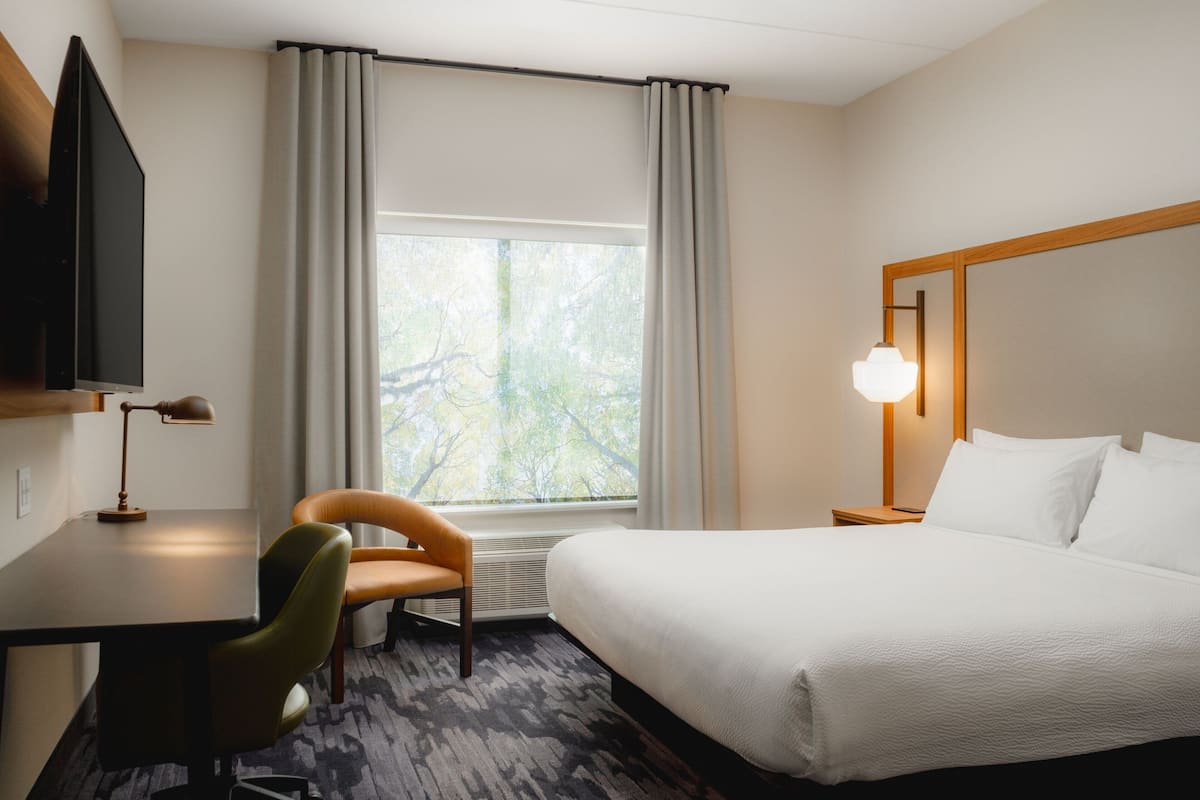 Une chambre d’hôtel comprenant un lit, un bureau, une chaise, un téléviseur, une fenêtre avec des rideaux et une table de chevet avec une lampe.