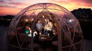 A transparent dome structure with a table and chairs inside, surrounded by a cityscape at sunset.