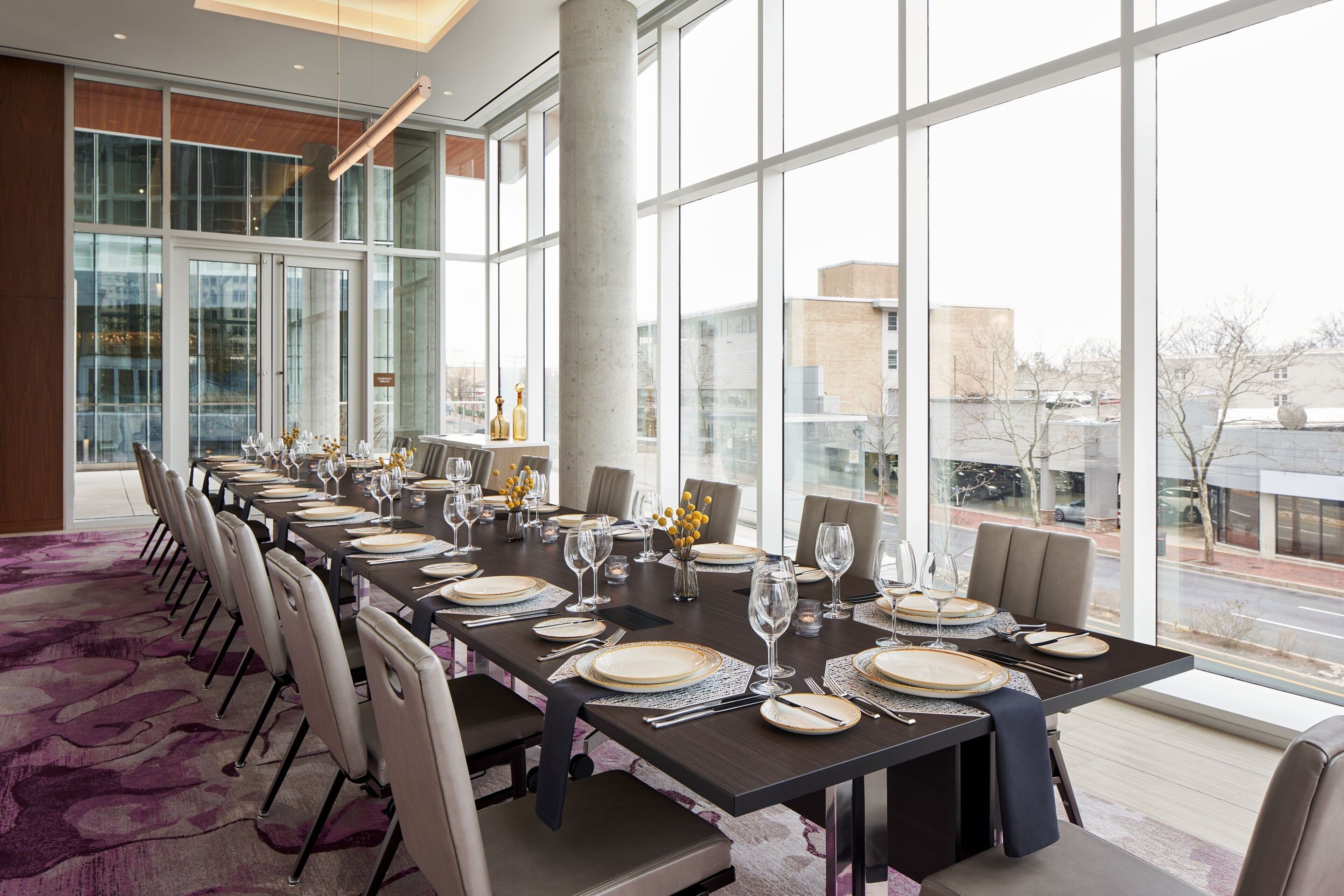 A long dining table set for a meal with multiple chairs, large windows, and a view of the cityscape.