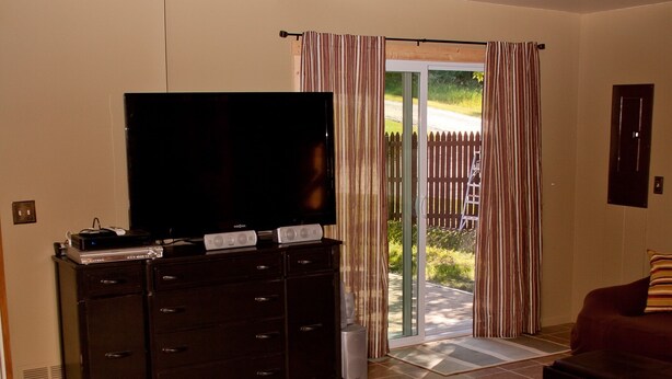 A living room with a flat-screen TV, dark wooden entertainment unit, brown leather sofa, and a sliding glass door leading to a patio. - Cozy, quirky and quaint updated Farm House surrounded by Sleeping Bear Dunes. (Maple City)