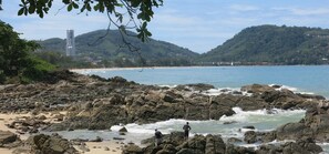 Une plage rocheuse où les vagues se brisent sur le rivage, un ciel dégagé et une vue lointaine sur un paysage urbain avec des bâtiments et des collines.