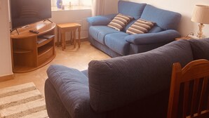 A living room with a blue sofa, a TV, a wooden entertainment unit, a small table, and a lamp. - Spacious 3 bed/2 bath house, on a gated complex, Fibre Wifi. Two communal pools. (Antigua)