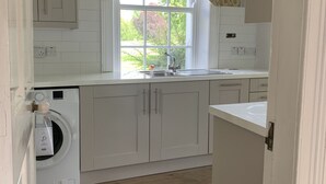 A kitchen with white cabinets, a built-in washing machine, a window with floral curtains, and a view of greenery outside.