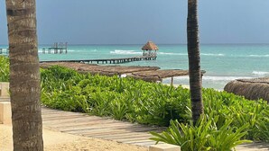 Vista de la playa con un muelle de madera que se adentra en el océano, una cabaña de techo de paja y abundante vegetación.