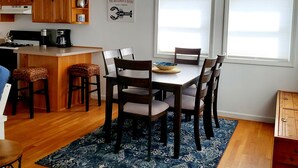 A modern kitchen with wooden cabinets, a dining area with a table and chairs, and a kitchen island with a coffee maker. - Steps to the Beach, Cozy Year Round Cottage 🐾 Pet Friendly (Old Saybrook)