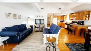 A modern living room with a blue sofa, a white armchair, and a TV. A kitchen with wooden cabinets and a bar area is visible in the background. - Steps to the Beach, Cozy Year Round Cottage 🐾 Pet Friendly (Old Saybrook)