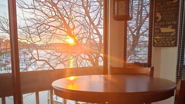 Ein Essbereich mit einem runden Tisch und Hocker, große Fenster mit Blick auf einen Sonnenuntergang über dem Wasser, und eine Wand mit einem Schild im Fischdesign.