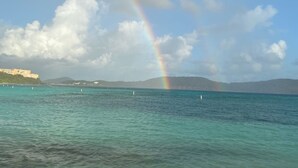 Klarer Blick auf eine Küstenlandschaft mit Regenbogen über dem Meer, einem entfernten Gebäude und einer hügeligen Landschaft.