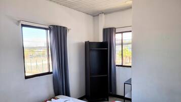 Une chambre avec deux lits, une étagère noire, une petite table et une fenêtre avec des rideaux.