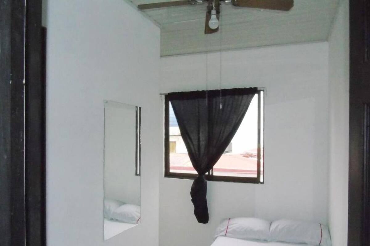 A bedroom with a bed, a window with a curtain, a ceiling fan, and a mirror on the wall.