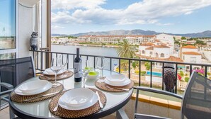 A balcony table set with plates, glasses, and a bottle, overlooking a coastal town with buildings and mountains.