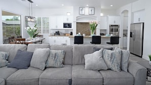 A modern living room with a grey sectional sofa, a dining area with a table and chairs, and a kitchen with white cabinets and stainless steel appliances. - Villa Serene Luxurious Resort Vacation Home - pool (Indio)