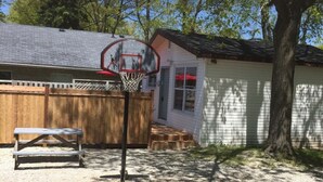 Een basketbalring in een achtertuin, met een houten bankje en een tuinhuisje op de achtergrond.