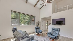 A spacious living room with a large window, two leather chairs, a ceiling fan, and a mounted TV. - Modern Alpine Cabin 3 Miles to Ski Cloudcroft (Cloudcroft)
