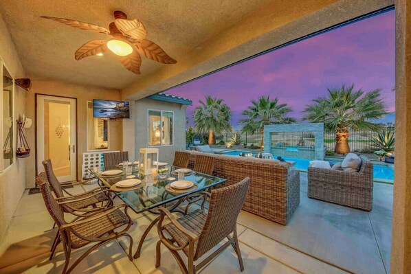 An outdoor dining area with wicker furniture, a glass table, and a view of a swimming pool and palm trees at dusk.