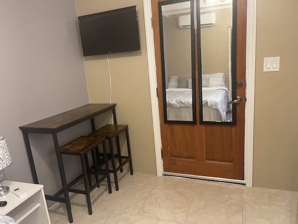 A small room with a wooden desk, two stools, a TV mounted on the wall, and a door with a mirror.