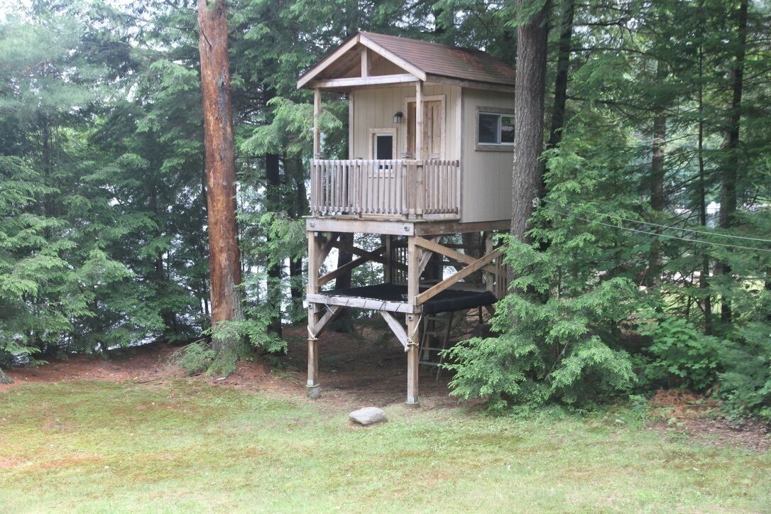 A treehouse with a wooden deck and a small porch, surrounded by dense forest.
