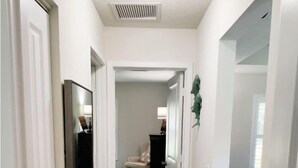 A narrow hallway with a light gray carpet, white walls, and a ceiling vent. There is a door on the left and a plant on the right. - Vero's Cove  (Vero Beach)