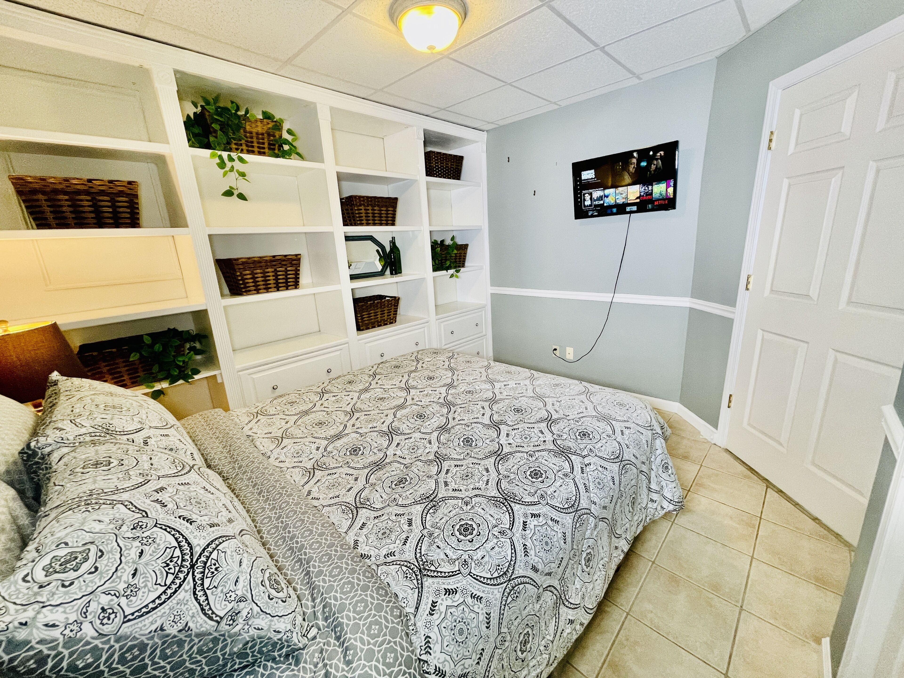 A bedroom with a bed, a TV mounted on the wall, and built-in shelving with baskets and plants.