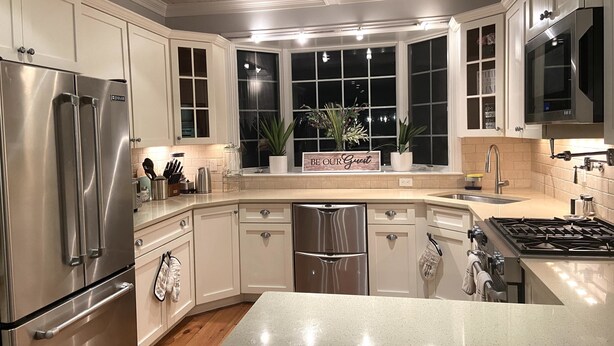 A modern kitchen with stainless steel appliances, white cabinetry, a built-in stove, and a window with a 'Be Our Guest' sign.