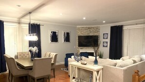 A modern living room with a dining area, featuring a sofa, chairs, a coffee table, and a TV mounted on the wall.