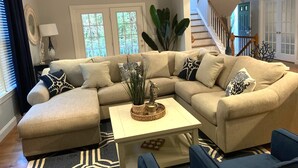 A living room with a beige sectional sofa, a white coffee table, and a blue armchair. There is a staircase in the background leading to an upper floor.
