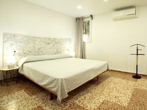 A bedroom with a bed, a nightstand, a lamp, a window with curtains, an air conditioning unit, and a floor with a speckled pattern. - Vacation home La Paz by Interhome (Calpe/Calp)