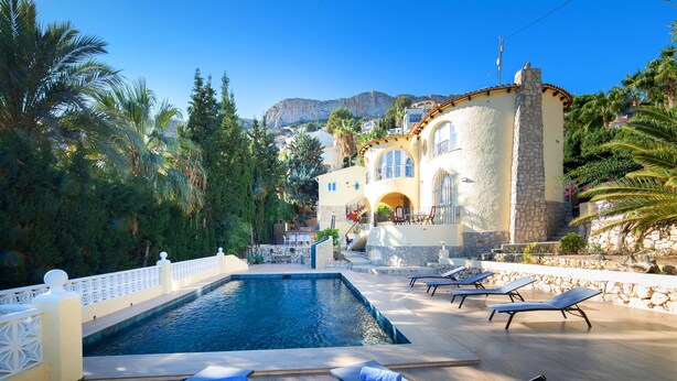 A pool area with lounge chairs, a stone wall, and a building with arched windows in the background. - Vacation home La Paz by Interhome (Calpe/Calp)