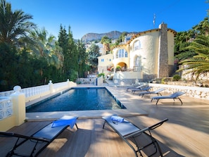 A pool area with lounge chairs, a stone wall, and a building with arched windows in the background. - Vacation home La Paz by Interhome (Calpe/Calp)