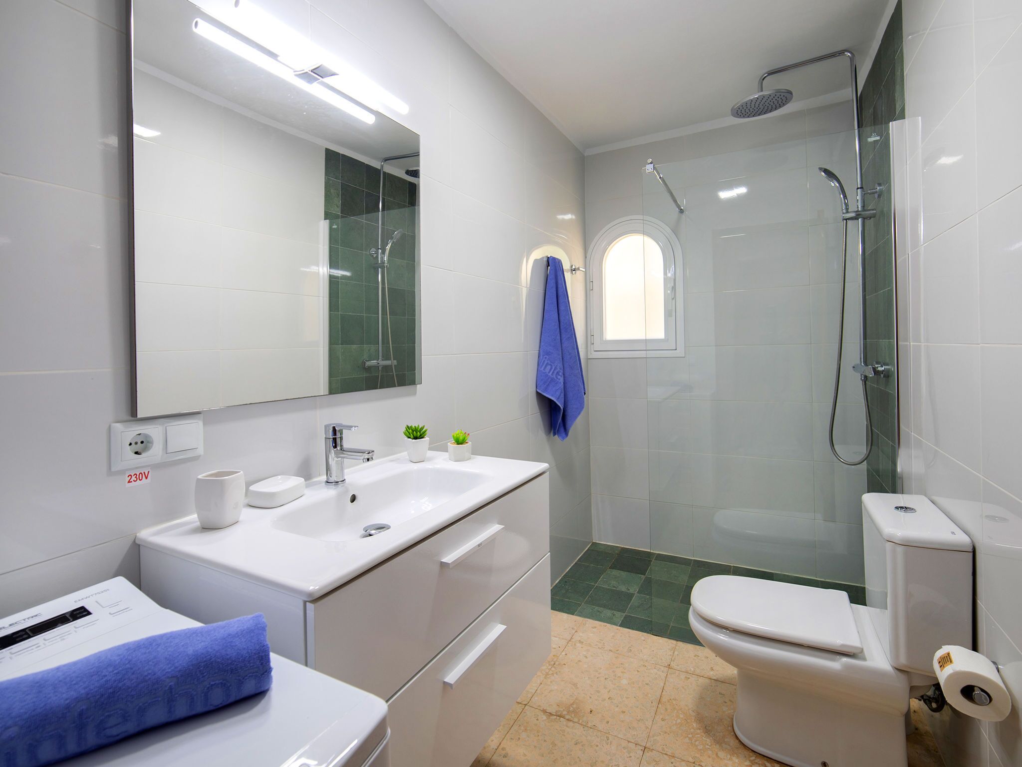 Ein modernes Badezimmer mit einem weißen Waschbecken, einem Spiegel, einer Toilette, einer Duschkabine und einem kleinen Fenster.