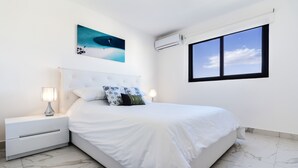 Une chambre à coucher avec un grand lit, du linge de lit blanc, une table de chevet avec une lampe, un climatiseur fixé au mur et une fenêtre donnant sur le ciel.