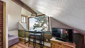 A cozy attic room with a wooden floor, a sofa, a dining table with stools, a TV on a wooden stand, and a large window offering a view of trees and water.