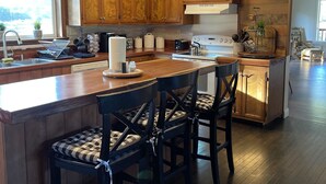 Cocina con gabinetes de madera, una isla central con taburetes de bar, un letrero que dice 'Eat' y un ventanal que proporciona luz natural.