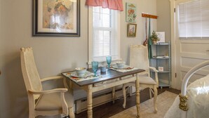 Un coin repas avec une table dressée pour deux personnes, deux chaises, un tableau encadré au mur, une fenêtre avec des stores et une porte avec une pancarte.