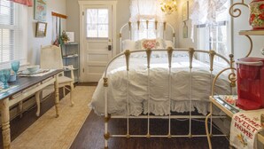 Une chambre avec un lit en métal blanc, un bureau en bois avec une chaise, une petite table sur laquelle est posée une plante en pot, et une fenêtre avec des rideaux.