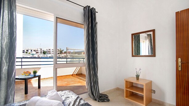 Una camera d'albergo con un letto, un tavolino con della frutta, vista su un edificio dalla finestra e una mensola in legno.